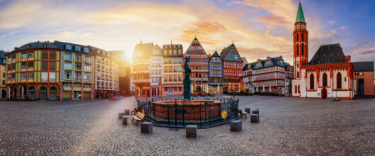 Een panoramisch stadsgezicht van het historische Römerberg-plein in Frankfurt, met traditionele vakwerkhuizen en een kerk rondom een centrale fontein bij zonsondergang.