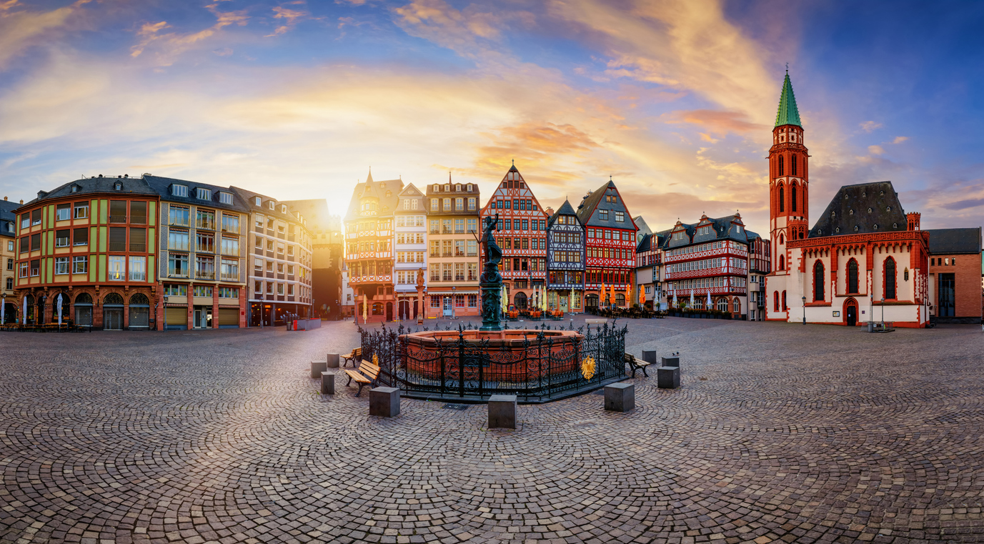 Een panoramisch stadsgezicht van het historische Römerberg-plein in Frankfurt, met traditionele vakwerkhuizen en een kerk rondom een centrale fontein bij zonsondergang.