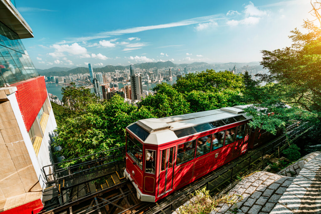 De rode Peak Tram-kabeltrein rijdt over een steile helling omhoog in Hongkong, met op de achtergrond een weids uitzicht over de skyline en de haven.
