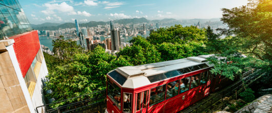 De rode Peak Tram-kabeltrein rijdt over een steile helling omhoog in Hongkong, met op de achtergrond een weids uitzicht over de skyline en de haven.