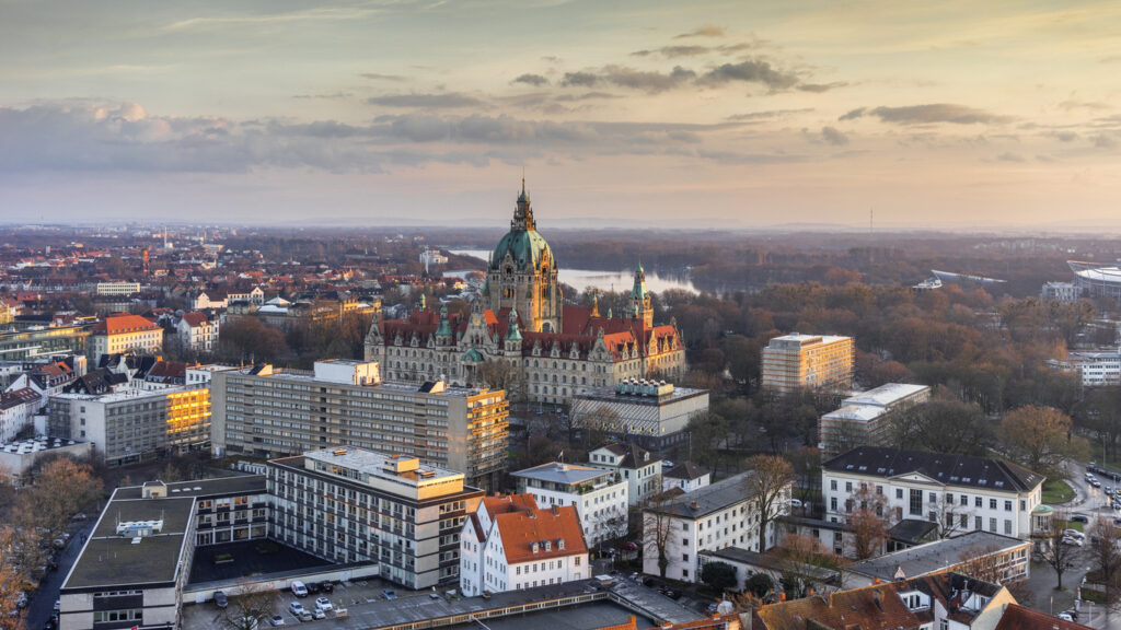 Een luchtfoto van de stad Hannover tijdens zonsondergang. In het midden staat een groot, historisch stadhuispaleis met een opvallend grote, groene koepel. Rondom het stadhuis liggen modernere flatgebouwen, veel bomen en op de achtergrond een meer onder een lichtbewolkte lucht.