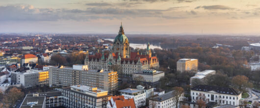 Een luchtfoto van de stad Hannover tijdens zonsondergang. In het midden staat een groot, historisch stadhuispaleis met een opvallend grote, groene koepel. Rondom het stadhuis liggen modernere flatgebouwen, veel bomen en op de achtergrond een meer onder een lichtbewolkte lucht.