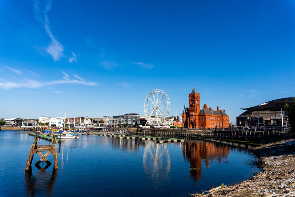 Uitzicht over de haven van Cardiff Bay onder een blauwe lucht met lichte wolken. Rechts staat een opvallend rood bakstenen gebouw met een klokkentoren en daarnaast staat een groot wit reuzenrad. Het water op de voorgrond is rustig en laat de spiegeling van de gebouwen zien.