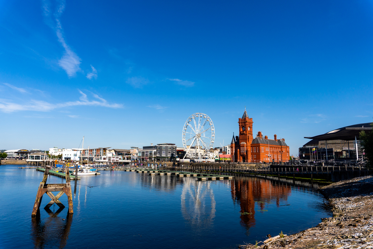 Uitzicht over de haven van Cardiff Bay onder een blauwe lucht met lichte wolken. Rechts staat een opvallend rood bakstenen gebouw met een klokkentoren en daarnaast staat een groot wit reuzenrad. Het water op de voorgrond is rustig en laat de spiegeling van de gebouwen zien.