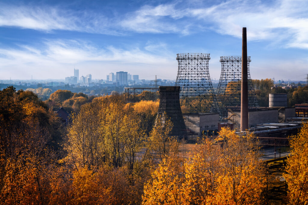 Uitzicht over een herfstlandschap met goudgele en oranje bomen op de voorgrond. Rechts staan de metalen constructies van oude industriële koeltorens en een hoge bakstenen schoorsteen van een fabriek. Helemaal op de achtergrond zie je de verre skyline van de stad onder een blauwe lucht met witte wolken.