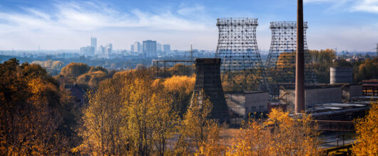 Uitzicht over een herfstlandschap met goudgele en oranje bomen op de voorgrond. Rechts staan de metalen constructies van oude industriële koeltorens en een hoge bakstenen schoorsteen van een fabriek. Helemaal op de achtergrond zie je de verre skyline van de stad onder een blauwe lucht met witte wolken.