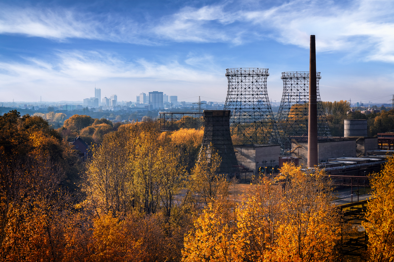 Uitzicht over een herfstlandschap met goudgele en oranje bomen op de voorgrond. Rechts staan de metalen constructies van oude industriële koeltorens en een hoge bakstenen schoorsteen van een fabriek. Helemaal op de achtergrond zie je de verre skyline van de stad onder een blauwe lucht met witte wolken.