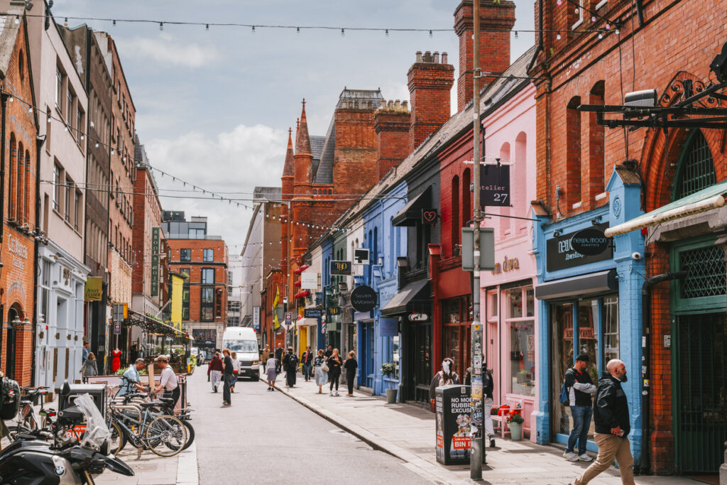 Een sfeervolle winkelstraat in Dublin met historische gebouwen van rode baksteen aan de linkerkant en vrolijk gekleurde winkelgevels in het blauw, roze en rood aan de rechterkant. Er wandelen verschillende mensen op de straat en op de stoep, en er hangen feestverlichtingssnoeren boven de weg.
