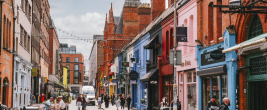Een sfeervolle winkelstraat in Dublin met historische gebouwen van rode baksteen aan de linkerkant en vrolijk gekleurde winkelgevels in het blauw, roze en rood aan de rechterkant. Er wandelen verschillende mensen op de straat en op de stoep, en er hangen feestverlichtingssnoeren boven de weg.