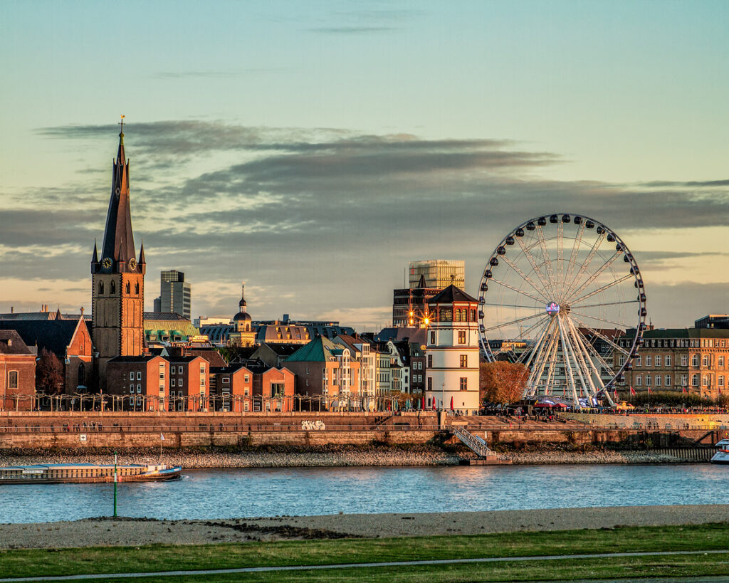 Een stadsgezicht van Düsseldorf vanaf de overkant van de rivier de Rijn tijdens de avondzon. Links staat een oude kerk met een opvallend gedraaide, donkere spits. In het midden staat een ronde, witte kasteeltoren en rechts staat een groot, verlicht reuzenrad tegen een bewolkte lucht.