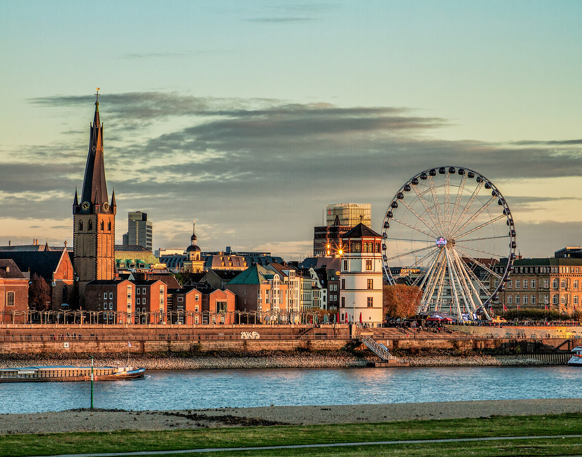 Een stadsgezicht van Düsseldorf vanaf de overkant van de rivier de Rijn tijdens de avondzon. Links staat een oude kerk met een opvallend gedraaide, donkere spits. In het midden staat een ronde, witte kasteeltoren en rechts staat een groot, verlicht reuzenrad tegen een bewolkte lucht.