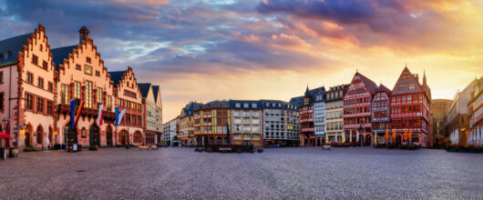 Een brede panoramafoto van een historisch plein met kasseien onder een kleurrijke avondlucht. Links staat het oude stadhuis met drie trapgevels en vlaggen aan de muur. Aan de rechterkant staat een rij traditionele, kleurrijke vakwerkhuizen.