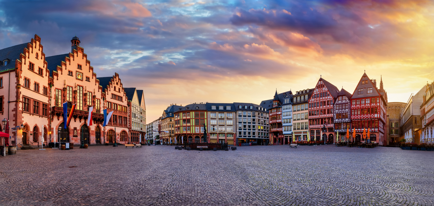Een brede panoramafoto van een historisch plein met kasseien onder een kleurrijke avondlucht. Links staat het oude stadhuis met drie trapgevels en vlaggen aan de muur. Aan de rechterkant staat een rij traditionele, kleurrijke vakwerkhuizen.