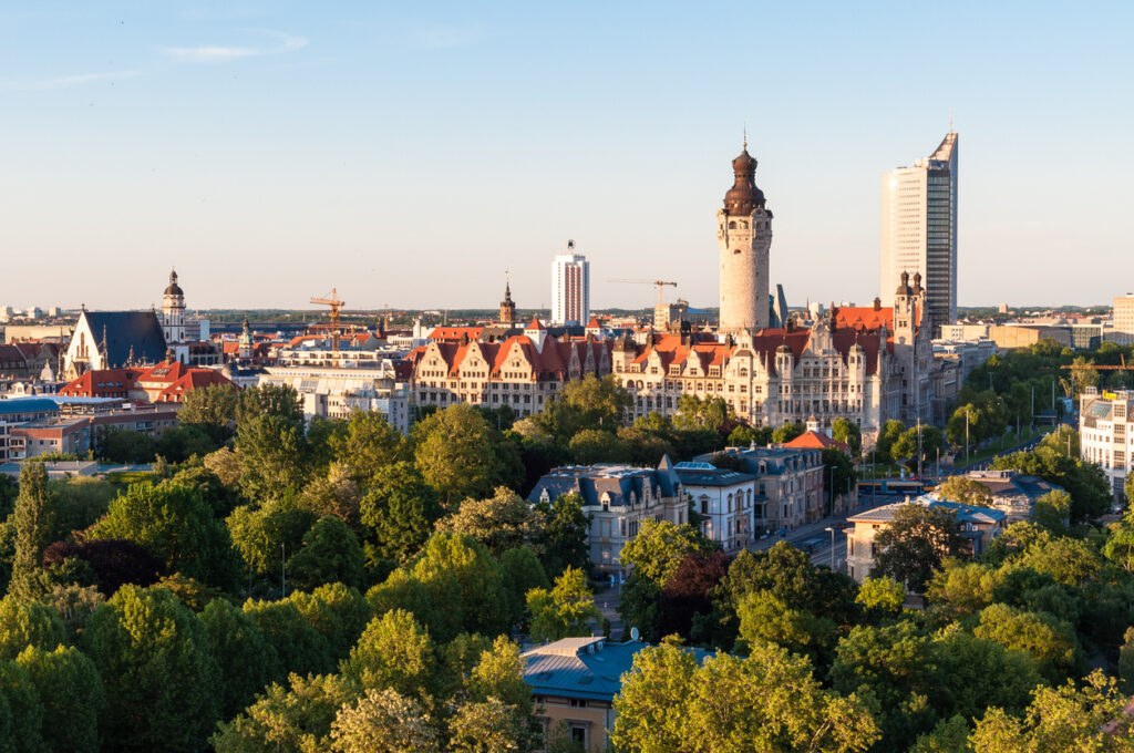 Een luchtfoto van een stad onder een heldere, blauwe lucht. Op de voorgrond staan veel groene bomen. Daarachter staat een groot, historisch stadhuis met een opvallend hoge, ronde toren. Rechts van het stadhuis staat een hele hoge, moderne, zilvergrijze wolkenkrabber.