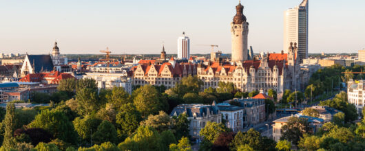 Een luchtfoto van een stad onder een heldere, blauwe lucht. Op de voorgrond staan veel groene bomen. Daarachter staat een groot, historisch stadhuis met een opvallend hoge, ronde toren. Rechts van het stadhuis staat een hele hoge, moderne, zilvergrijze wolkenkrabber.