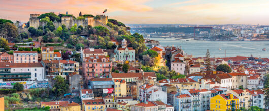 Uitzicht over de daken van een stad op een heuvel onder een zonsondergang met roze en blauwe wolken. Bovenop de heuvel links staat een oud stenen kasteel met vlaggen. De huizen eronder hebben oranjerode daken en witte of gekleurde muren. Rechts is een brede rivier te zien.