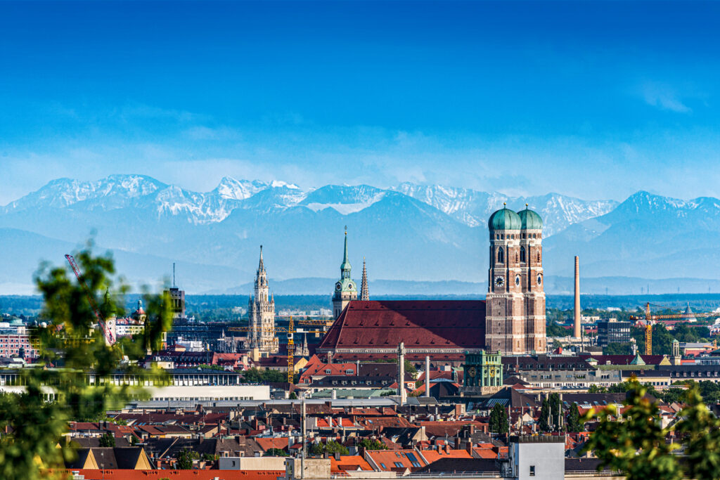 Een stadsgezicht van München onder een strakke blauwe lucht. In het midden valt de grote Frauenkirche op met haar twee kenmerkende torens met groene koepels. Op de achtergrond is een grote bergketen met besneeuwde toppen te zien. Op de voorgrond zijn groene bladeren en de daken van de stad zichtbaar.