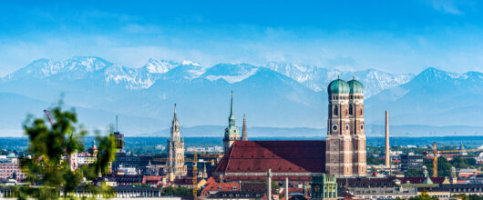 Een stadsgezicht van München onder een strakke blauwe lucht. In het midden valt de grote Frauenkirche op met haar twee kenmerkende torens met groene koepels. Op de achtergrond is een grote bergketen met besneeuwde toppen te zien. Op de voorgrond zijn groene bladeren en de daken van de stad zichtbaar.