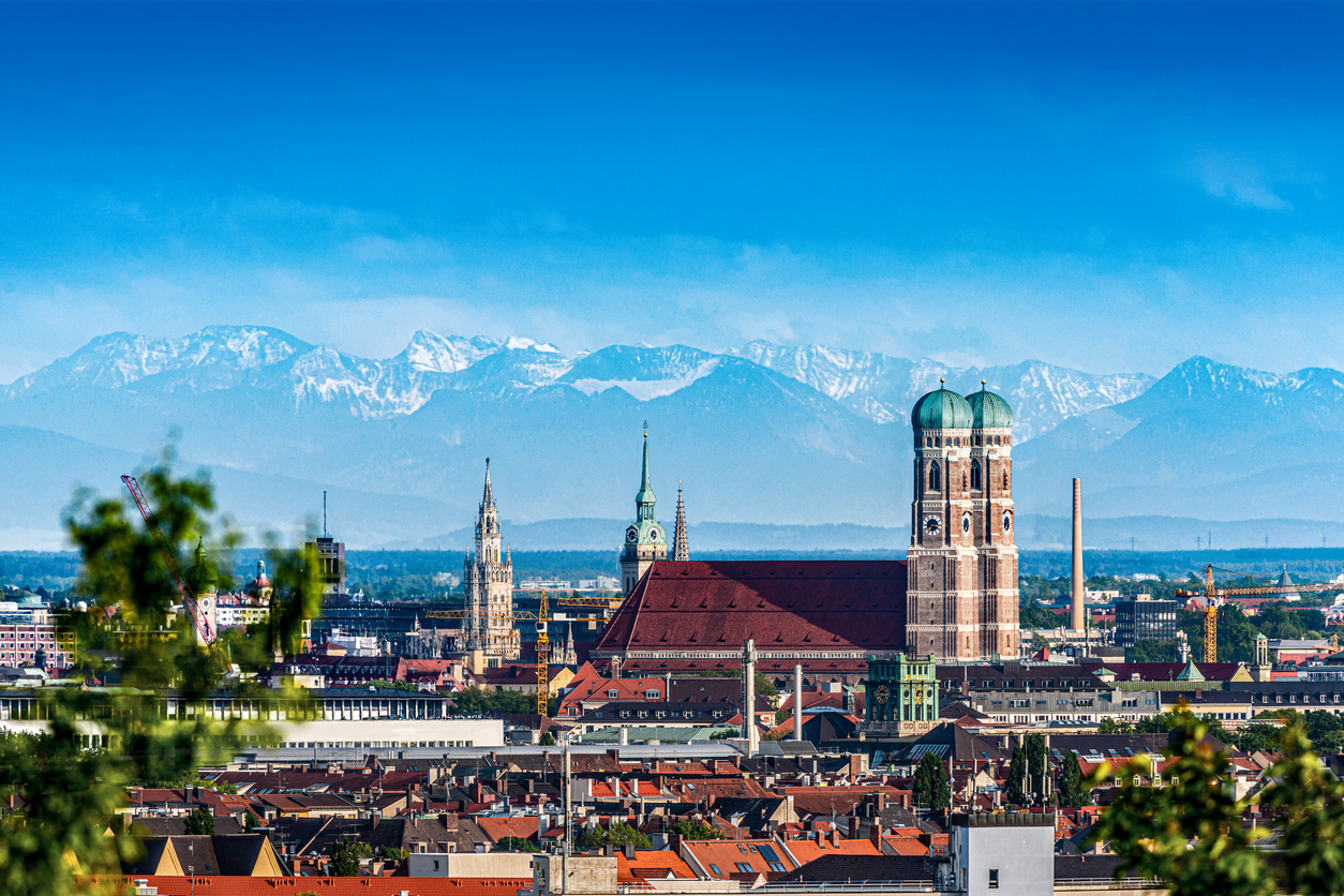 Een stadsgezicht van München onder een strakke blauwe lucht. In het midden valt de grote Frauenkirche op met haar twee kenmerkende torens met groene koepels. Op de achtergrond is een grote bergketen met besneeuwde toppen te zien. Op de voorgrond zijn groene bladeren en de daken van de stad zichtbaar.