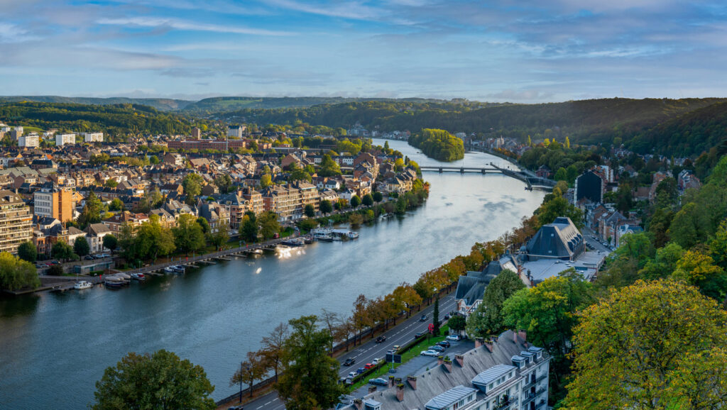 Een hoog panoramisch uitzicht over de rivier de Maas die door de stad Namen kronkelt, geflankeerd door huizen, wegen en groen begroeide heuvels.