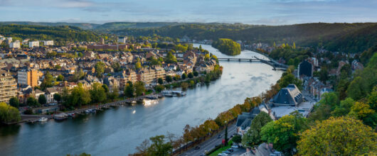 Een hoog panoramisch uitzicht over de rivier de Maas die door de stad Namen kronkelt, geflankeerd door huizen, wegen en groen begroeide heuvels.