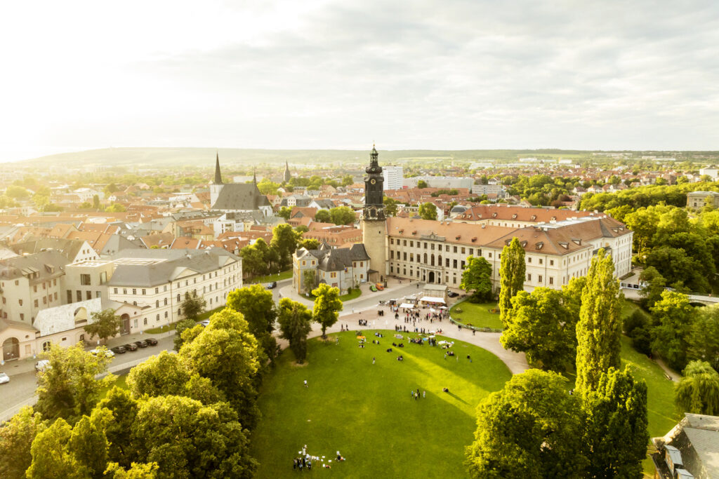 Luchtfoto in warm zonlicht van het barokke Stadtslot Weimar (Duitsland) met zijn opvallende kasteeltoren, omringd door een groen grasveld met mensen en de historische binnenstad op de achtergrond.