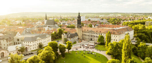 Luchtfoto in warm zonlicht van het barokke Stadtslot Weimar (Duitsland) met zijn opvallende kasteeltoren, omringd door een groen grasveld met mensen en de historische binnenstad op de achtergrond.