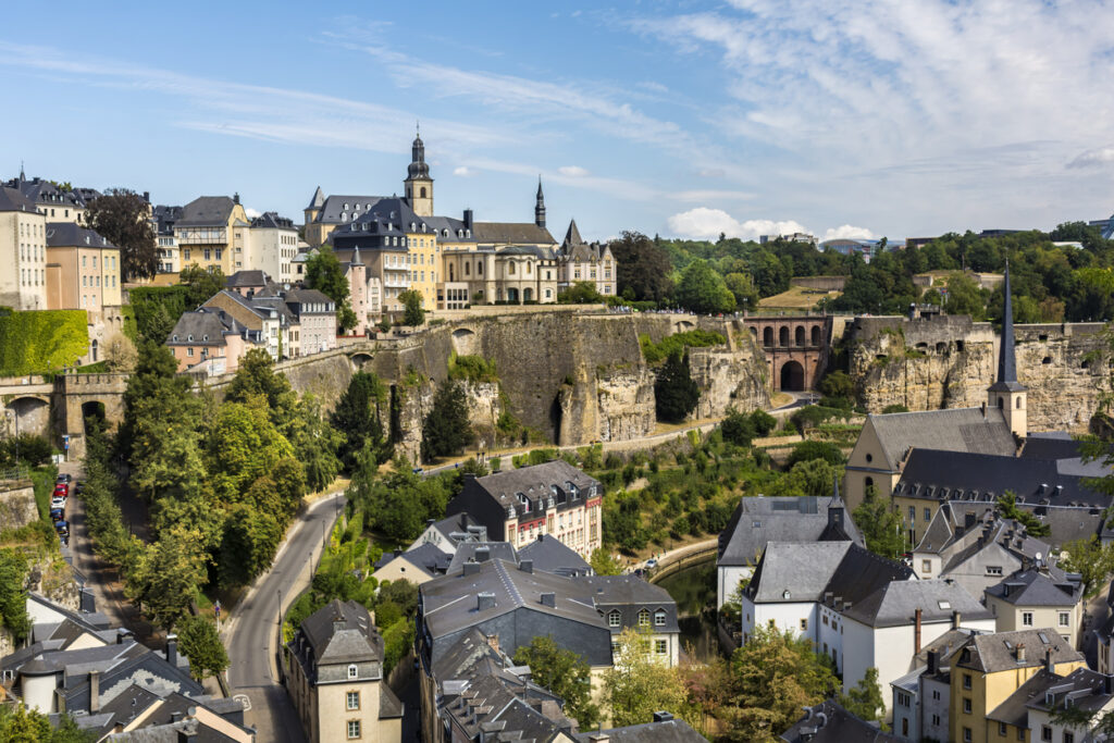 Een stadsgezicht van Luxemburg-Stad vanaf een hoog standpunt. In het dal liggen huizen met donkere daken en een kerk met een spitse toren langs een riviertje. Daarachter rijzen hoge, steile rotswanden op met oude vestingmuren en grote, historische gebouwen op de top, onder een lichtbewolkte blauwe lucht.