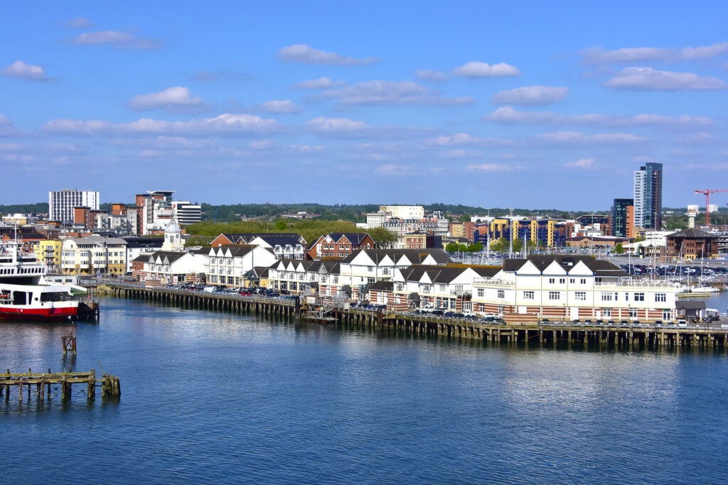 Blik vanaf het water op een lange houten pier in Southampton met een rij witte gebouwen met donkere daken erop. Links ligt een rood-witte veerboot aangemeerd. Op de achtergrond is de skyline van de stad te zien met moderne flats onder een helderblauwe lucht met plukjes witte wolken.