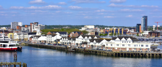 Blik vanaf het water op een lange houten pier in Southampton met een rij witte gebouwen met donkere daken erop. Links ligt een rood-witte veerboot aangemeerd. Op de achtergrond is de skyline van de stad te zien met moderne flats onder een helderblauwe lucht met plukjes witte wolken.