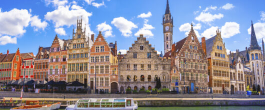 Kleurrijke historische gildehuizen aan de Graslei in Gent, weerspiegeld in het water onder een blauwe lucht met wolken.