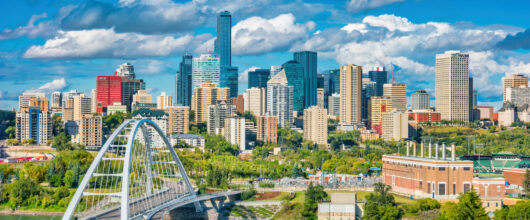 Een zonnig stadsgezicht op de skyline van Edmonton met moderne wolkenkrabbers en de opvallende witte Walterdale-boogbrug over de rivier.