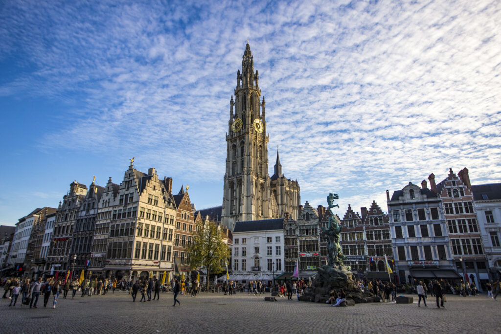 Uitzicht over een groot, druk stadsplein met kasseien onder een blauwe lucht vol kleine, witte wolken. Op de achtergrond steekt een enorme, rijk versierde gotische kerktoren met een gouden klok hoog boven de stad uit. Rondom het plein staan historische gildehuizen met trapgevels en rechts staat een groot standbeeld op een fontein.