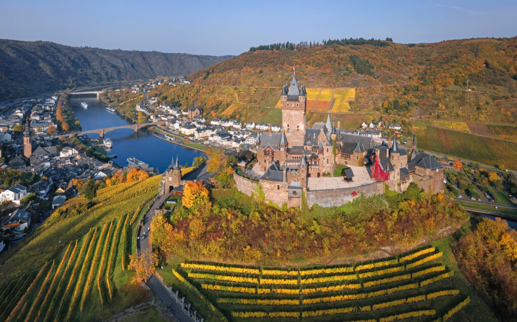 Een middeleeuws kasteel op een heuveltop omgeven door goudgele wijngaarden in de herfst. Op de achtergrond stroomt een rivier langs een Duits dorpje tussen beboste heuvels.