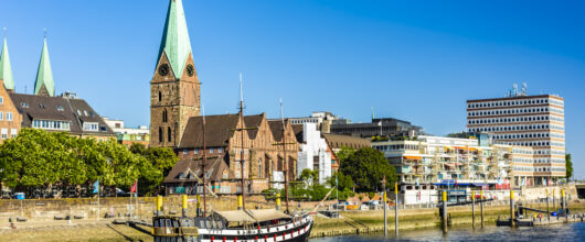 Een zonnig stadsgezicht in Bremen vanaf de rivier de Weser. Op de voorgrond ligt een zwart-wit historisch zeilschip aangemeerd aan de kade. Achter de kade staat de bakstenen St. Martinikirche met een hoge, groene torenspits. Aan de rechterkant strekt de Schlachte-promenade zich uit met moderne gebouwen en terrasjes onder een strakke, blauwe lucht.