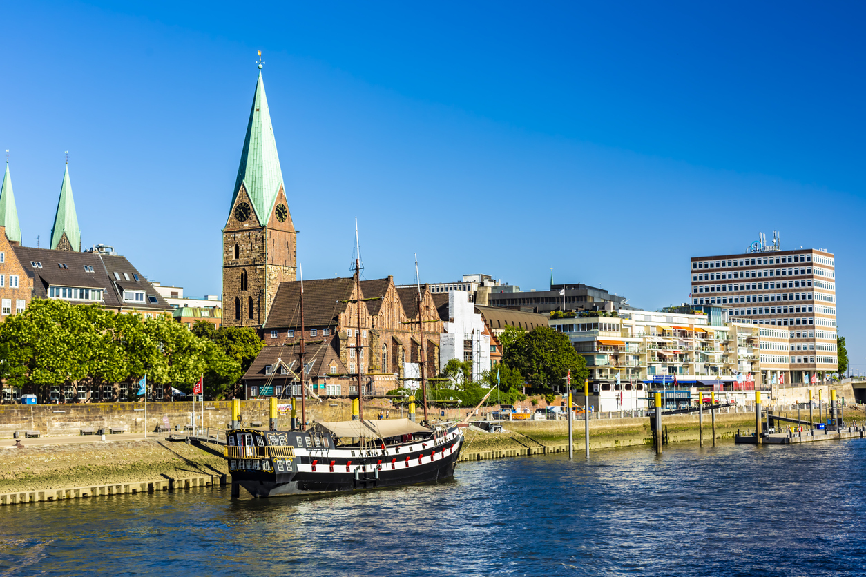 Een zonnig stadsgezicht in Bremen vanaf de rivier de Weser. Op de voorgrond ligt een zwart-wit historisch zeilschip aangemeerd aan de kade. Achter de kade staat de bakstenen St. Martinikirche met een hoge, groene torenspits. Aan de rechterkant strekt de Schlachte-promenade zich uit met moderne gebouwen en terrasjes onder een strakke, blauwe lucht.