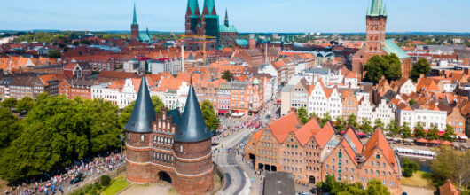 Een luchtfoto van de stad Lübeck onder een strakke, blauwe lucht met een paar witte wolken. Links op de voorgrond staat een grote, middeleeuwse stadspoort van rode baksteen met twee ronde torens en zwarte, spitse daken. Rechts daarvan staan oude pakhuizen en op de achtergrond zie je de rest van de stad met de hoge kerktorens.