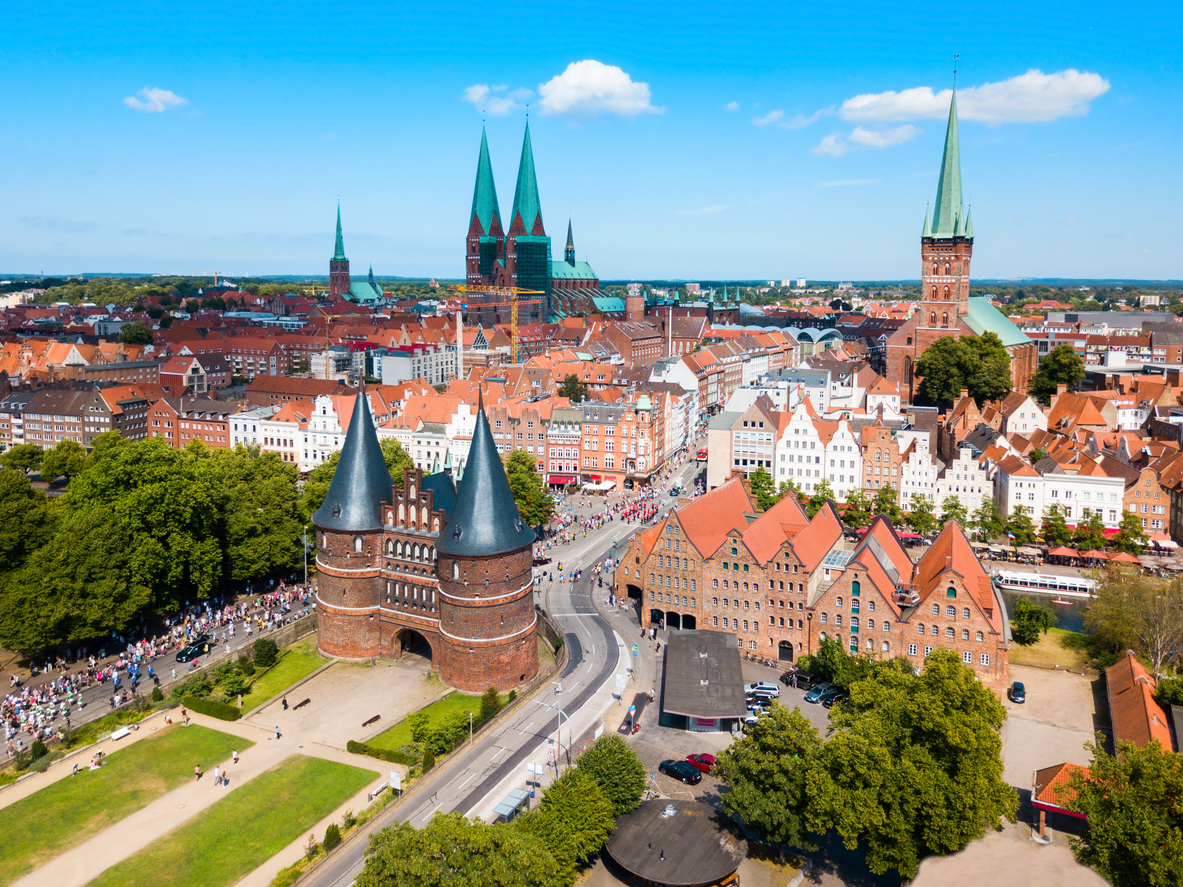 Een luchtfoto van de stad Lübeck onder een strakke, blauwe lucht met een paar witte wolken. Links op de voorgrond staat een grote, middeleeuwse stadspoort van rode baksteen met twee ronde torens en zwarte, spitse daken. Rechts daarvan staan oude pakhuizen en op de achtergrond zie je de rest van de stad met de hoge kerktorens.