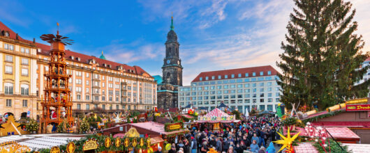 Een drukbezochte kerstmarkt in Dresden met verlichte houten kraampjes, een grote houten kerstpiramide en een historische kerktoren op de achtergrond.