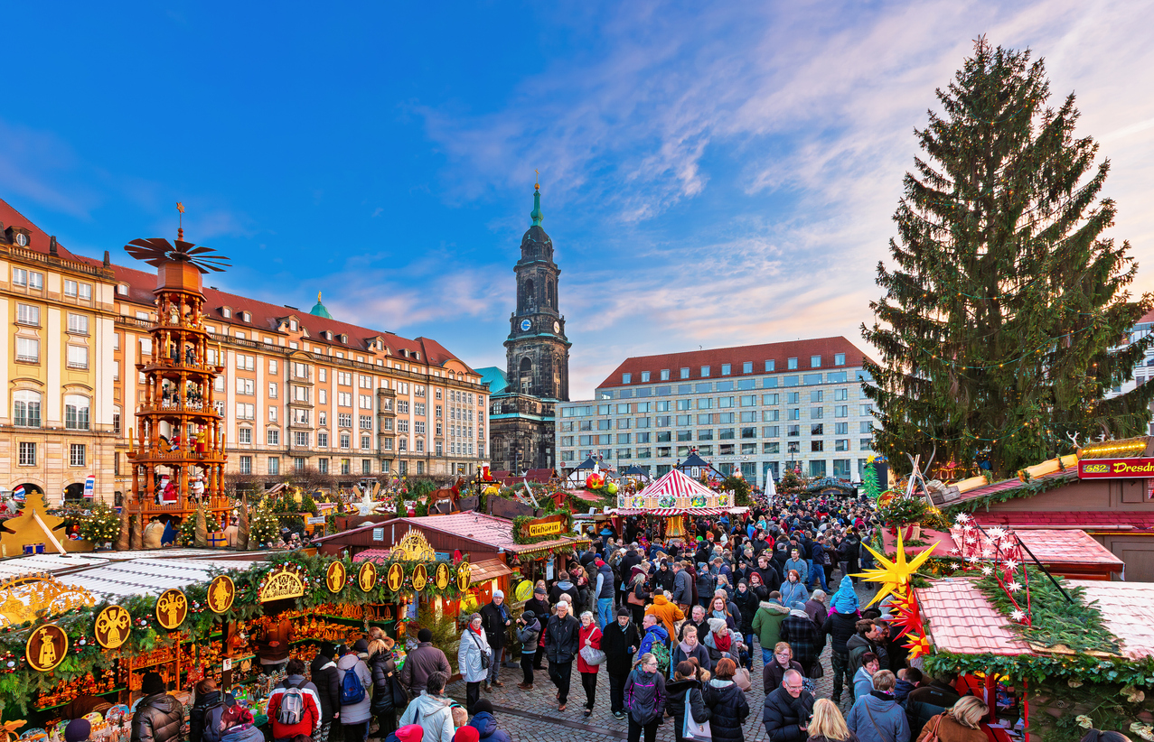 Een drukbezochte kerstmarkt in Dresden met verlichte houten kraampjes, een grote houten kerstpiramide en een historische kerktoren op de achtergrond.