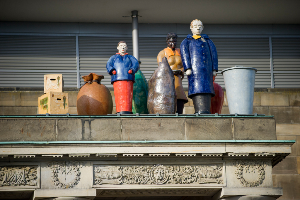 Een rij kleurrijke, geglazuurde keramische beelden van menselijke figuren met tassen en vaten, geplaatst op de rijk versierde historische daklijst van het Rote Palais in Kassel. Het is het documenta-kunstwerk 'Die Fremden' van Thomas Schütte.