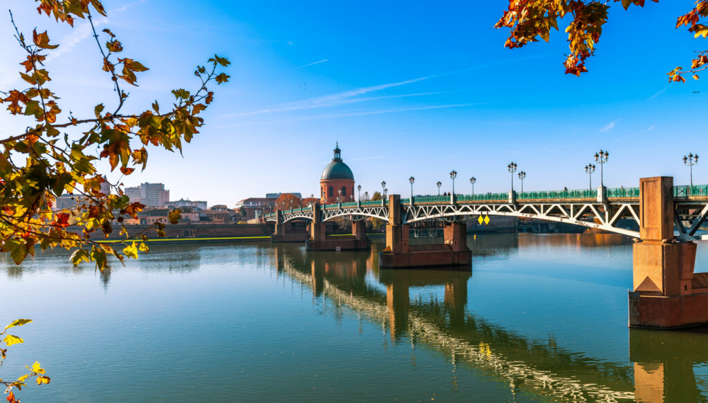 Uitzicht op een brede rivier op een zonnige dag. Een grote brug met metalen bogen steekt het water over en weerspiegelt in het oppervlak. Aan het einde van de brug staat een historisch gebouw met een grote, ronde koepel. Links en bovenin het beeld hangen herfstbladeren aan takken tegen een strakke, blauwe lucht.