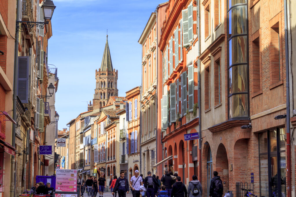 Een levendige, smalle straat in Toulouse met wandelaars tussen hoge, warmgekleurde gebouwen van rode baksteen en terracotta. Aan het einde van de straat rijst een grote, meerlaagse romaanse kerktoren met een spitse naald omhoog tegen een helderblauwe lucht.
