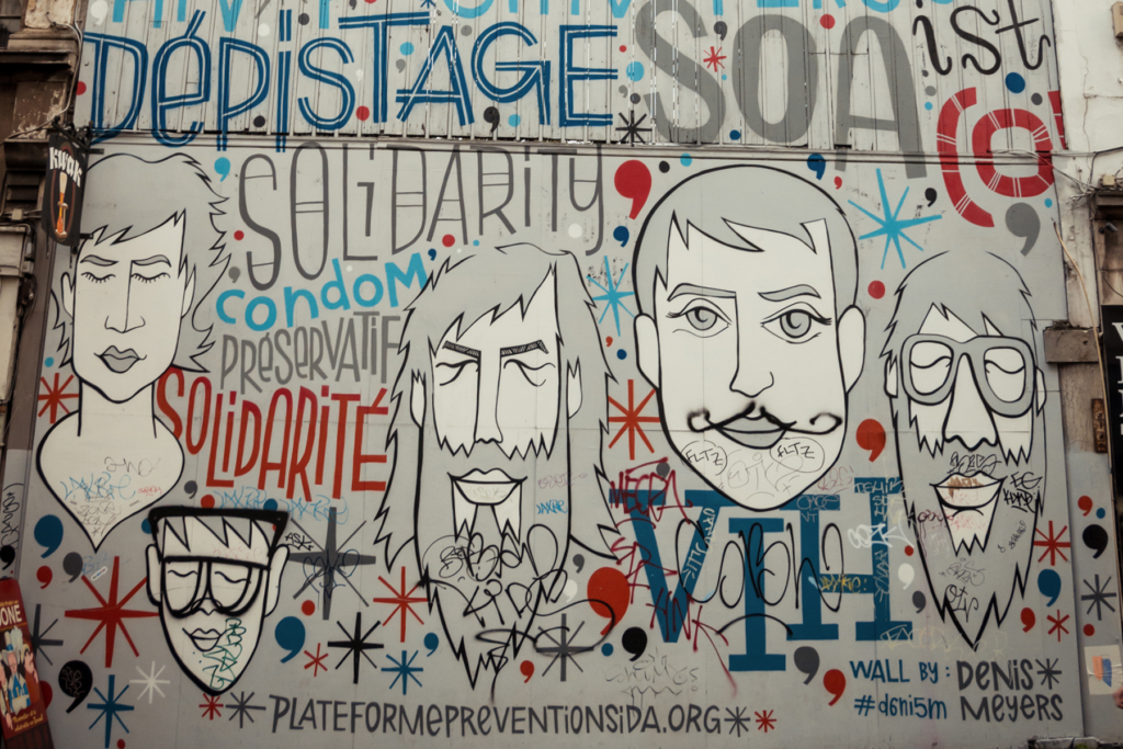 Grote street art mural met illustraties, teksten en rode linten over HIV, aids, solidariteit en preventie op een gevel in de stad.